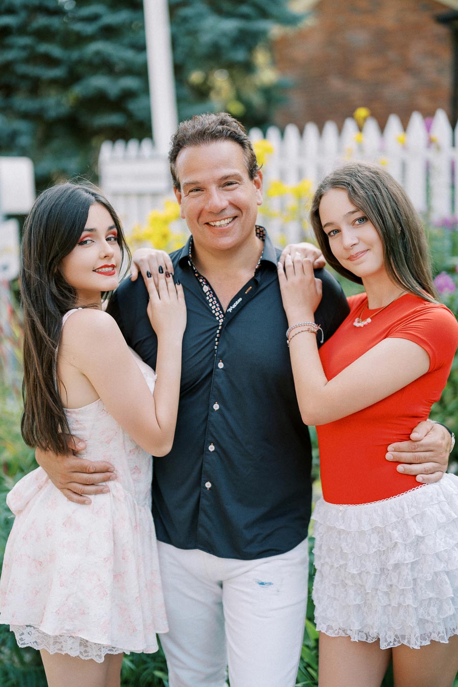 A cheerful man in a black shirt stands between two smiling women in a garden setting with white picket fences and blooming flowers in the background. The woman on the left wears a white floral dress, while the woman on the right wears a red top and white lace skirt.