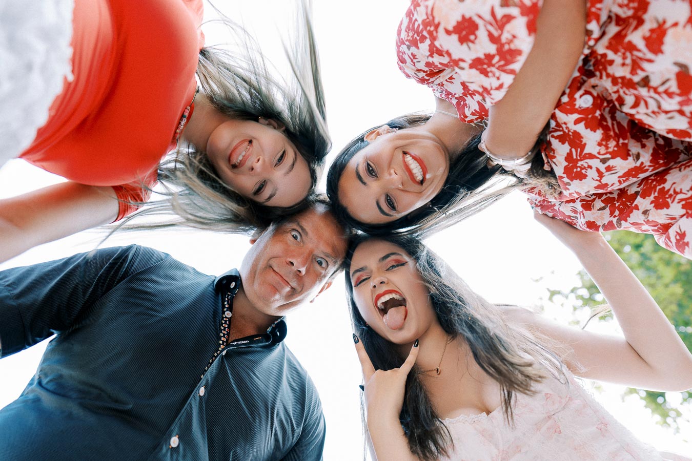 Group of four people happily looking down at camera, displaying playful expressions and wearing colorful clothing, shot from a low angle outdoors.
