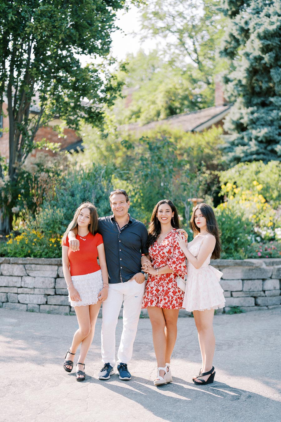 Family posing outdoors in a sunny garden, surrounded by lush greenery and a brick wall backdrop. Three women and one man, all smiling and dressed in summer attire.