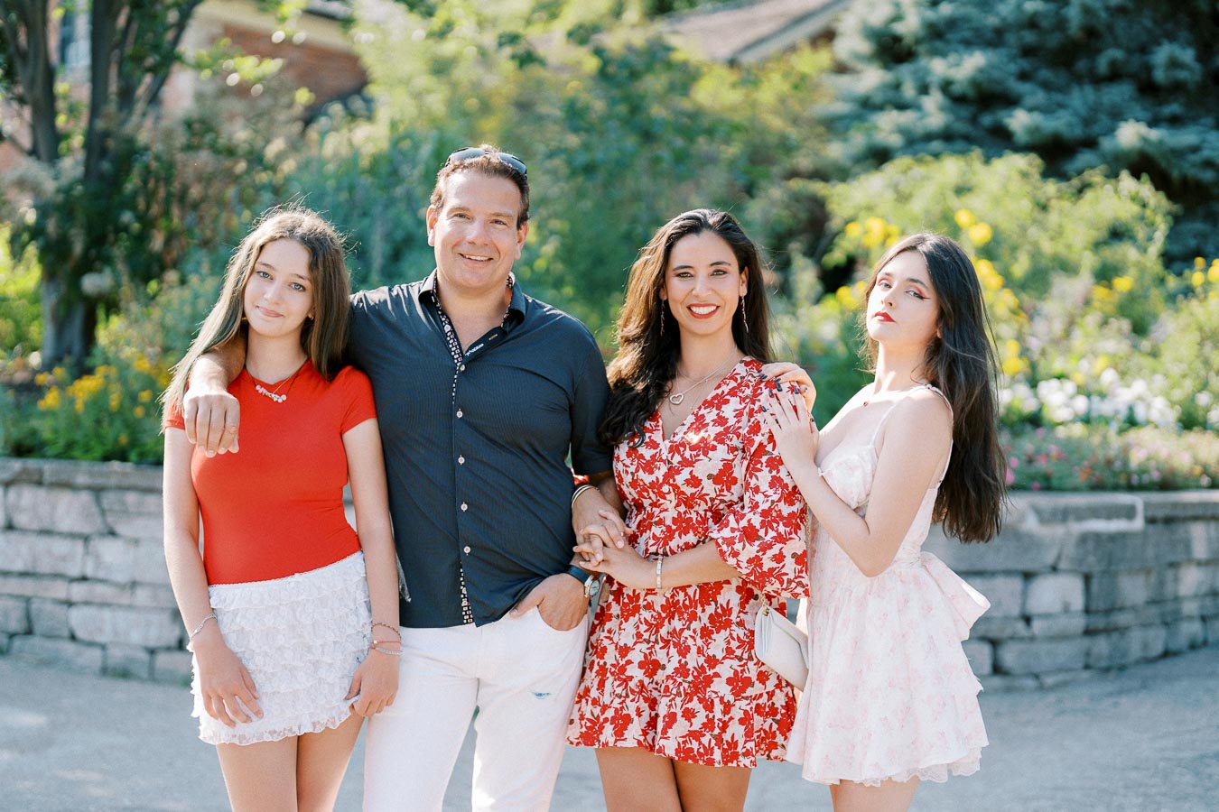 A family of four posing outdoors in a garden setting, with lush greenery and colorful flowers in the background, showcasing a cheerful and vibrant atmosphere.