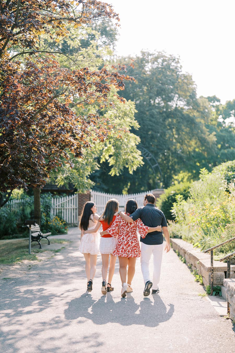 A group of four friends walking arm in arm down a park path, surrounded by lush green trees and a white picket fence, enjoying a sunny day.