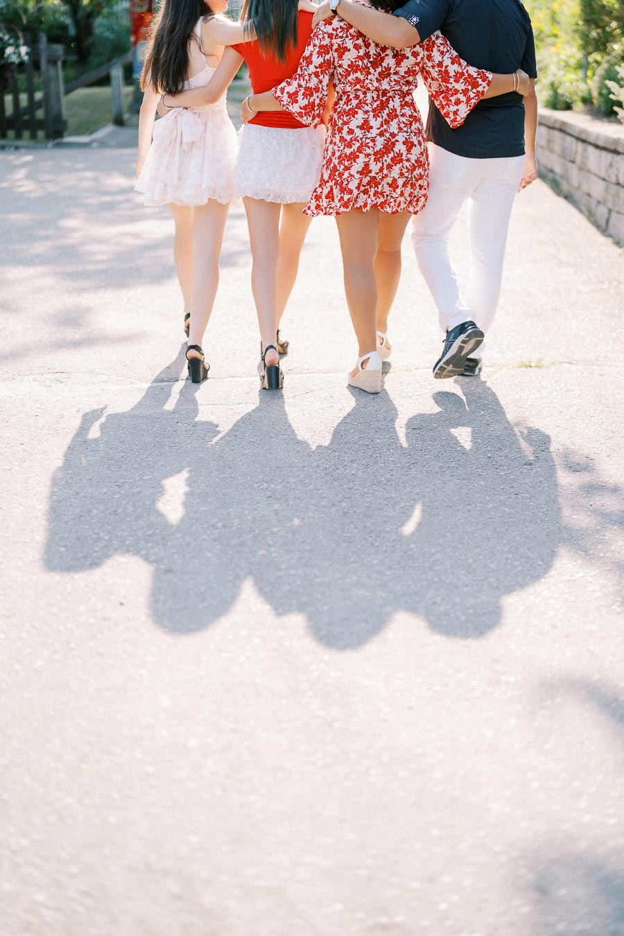 Four people walking arm in arm down a sunny path, casting long shadows on the pavement. They are wearing summer outfits, including dresses and casual attire, emphasizing a sense of friendship and togetherness.