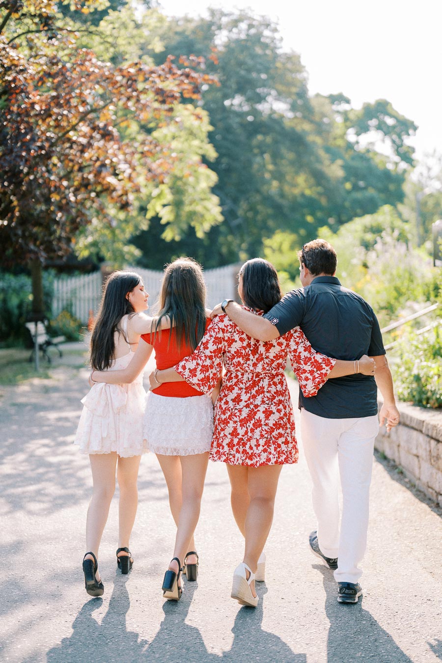 Four people walking arm-in-arm on a sunny park pathway, surrounded by lush greenery, expressing friendship and joy.