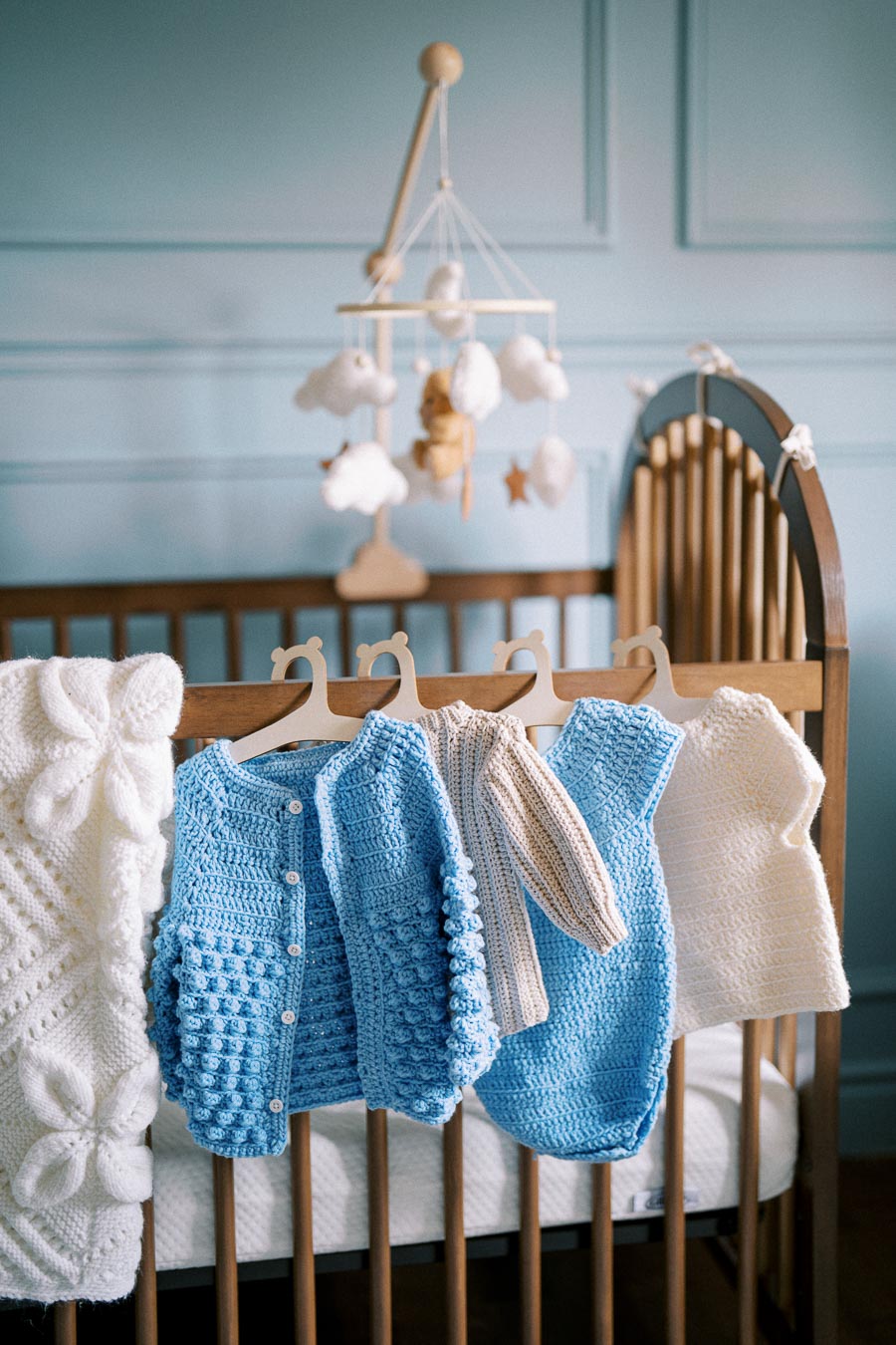 Crochet baby sweaters hanging on a crib in a nursery, with a mobile above.
