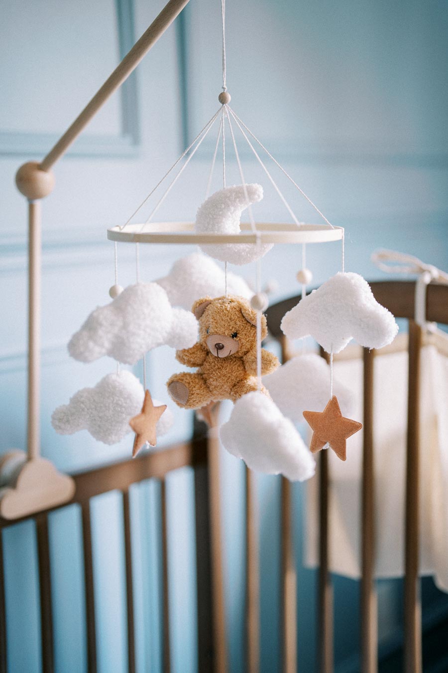 Adorable baby crib mobile featuring a plush teddy bear with fluffy white clouds and star accents, hanging over a wooden crib in a soothing nursery setting.