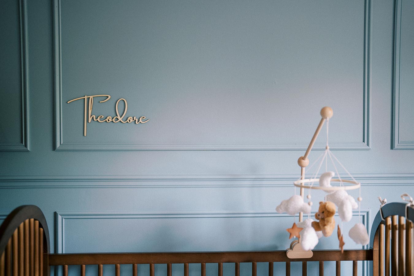 Blue nursery room with personalized name Theodore on the wall, featuring a wooden crib and a mobile with clouds and stars.