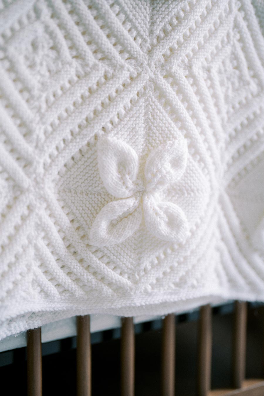 Close-up of a detailed knitted white blanket with a raised floral pattern, displayed on a wooden crib. Perfect for home decor or baby nurseries.