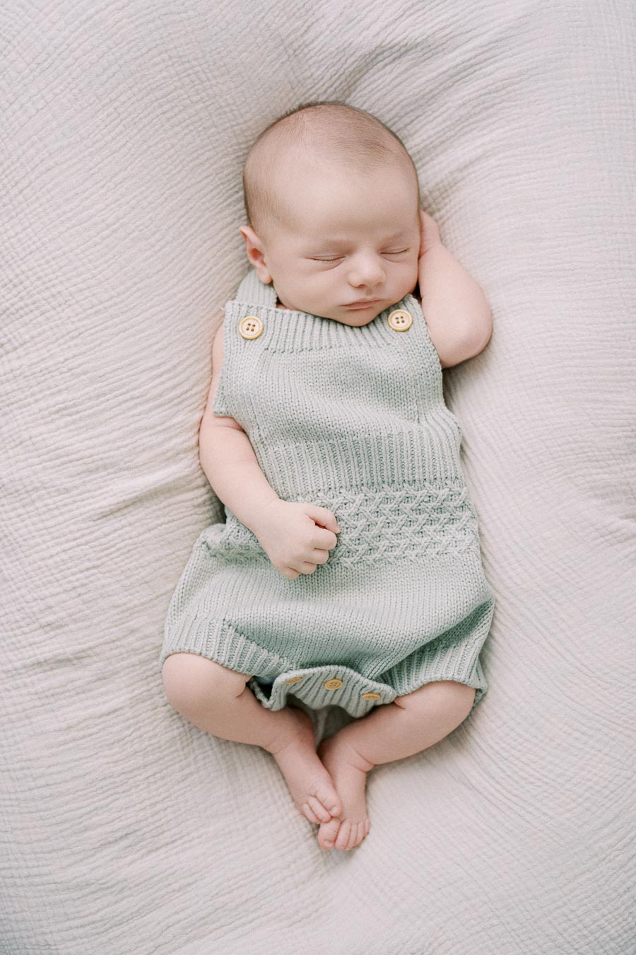 Cute sleeping baby wearing a knitted green romper with button details, resting peacefully on a soft, textured blanket.