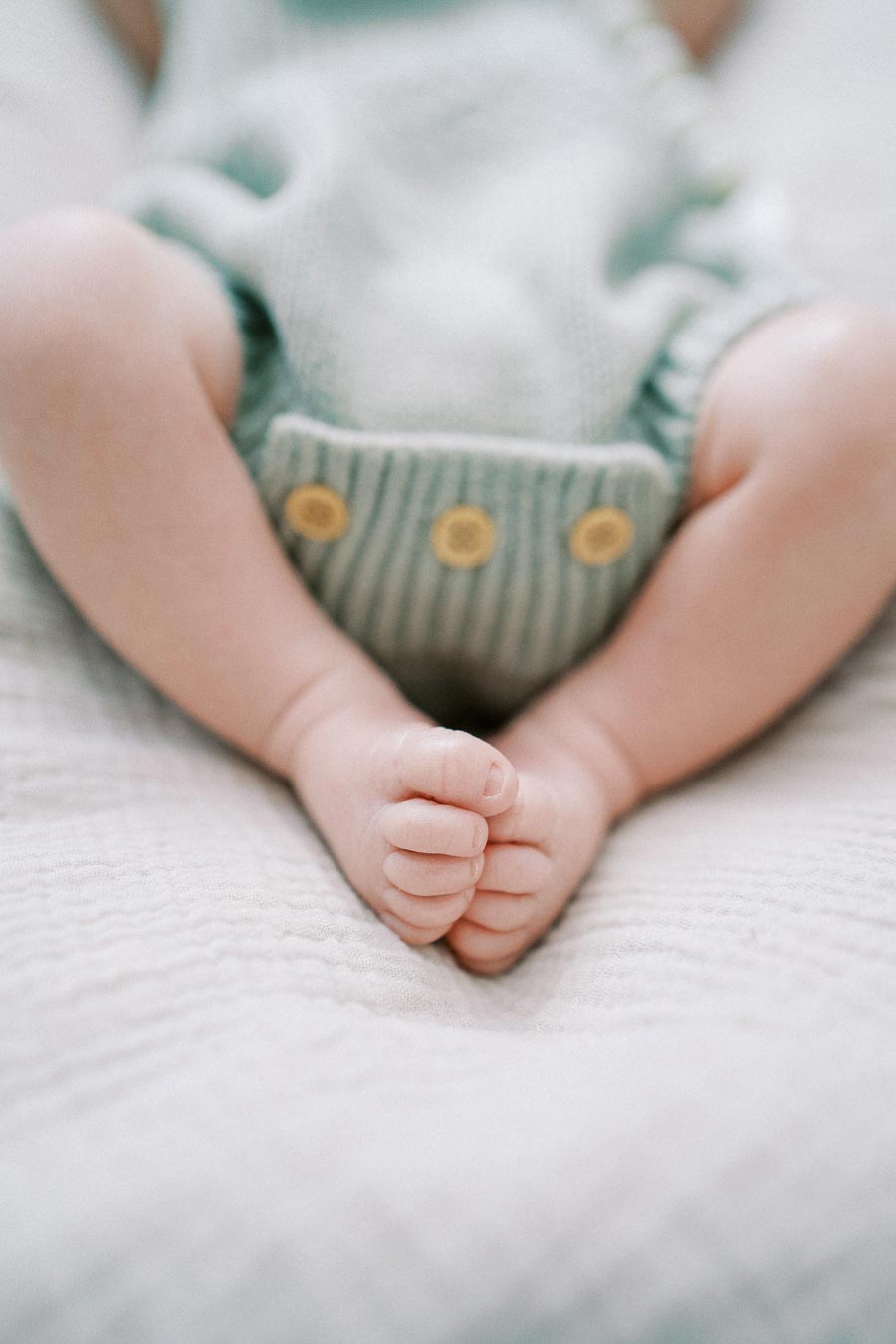 Baby wearing a mint green knitted romper with yellow buttons, showing small bare feet on a soft, textured white blanket.