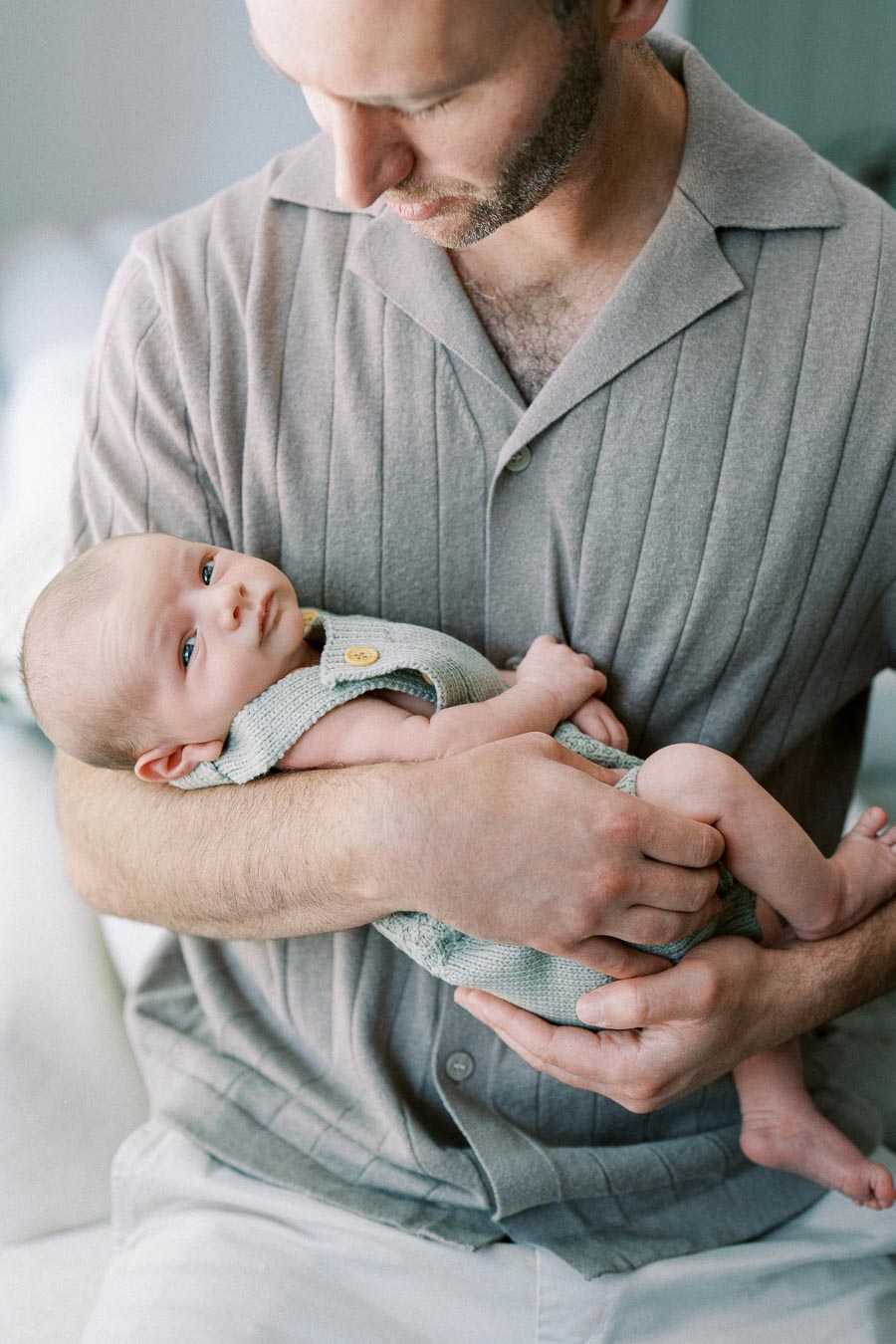 A father gently cradles his newborn baby in his arms, showcasing a tender moment of bonding and care, both dressed in soft, neutral-toned clothing.