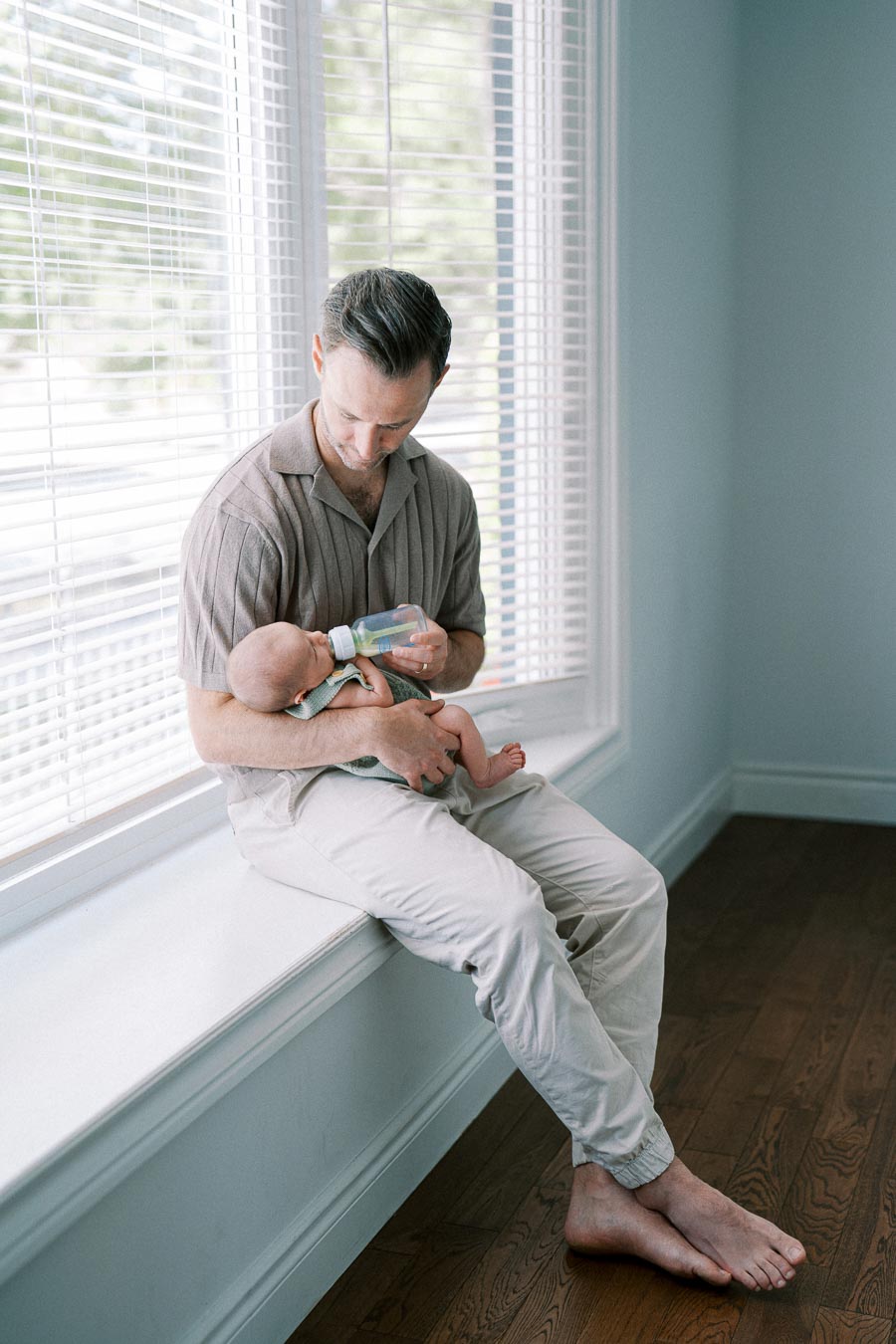 A father sitting on a windowsill, gently feeding his baby with a bottle, in a softly lit room with wooden floors and bright window blinds.