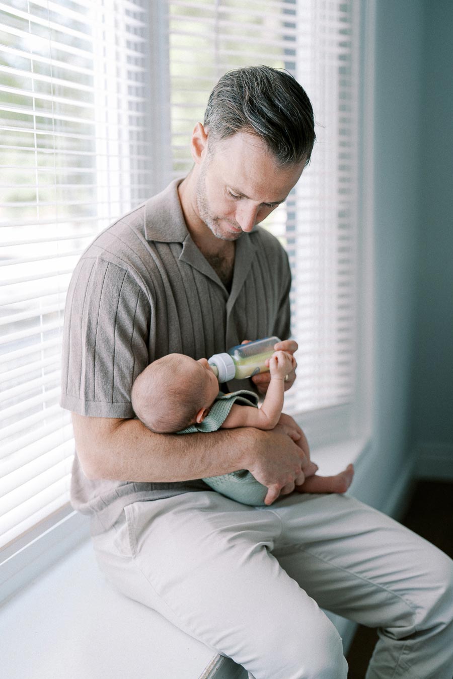 Father feeding baby with bottle while sitting by window blinds, showcasing a tender parenting moment.