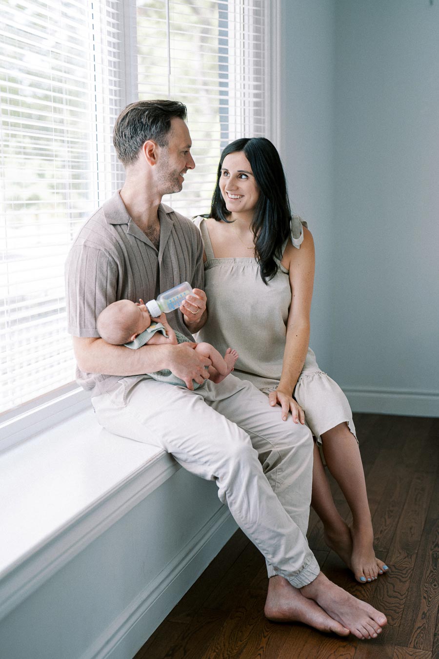 A couple sitting by a window feeding a baby with a bottle. The man holds the baby while the woman, wearing a beige dress, looks at him smiling. They are in a softly lit room with hardwood floors.
