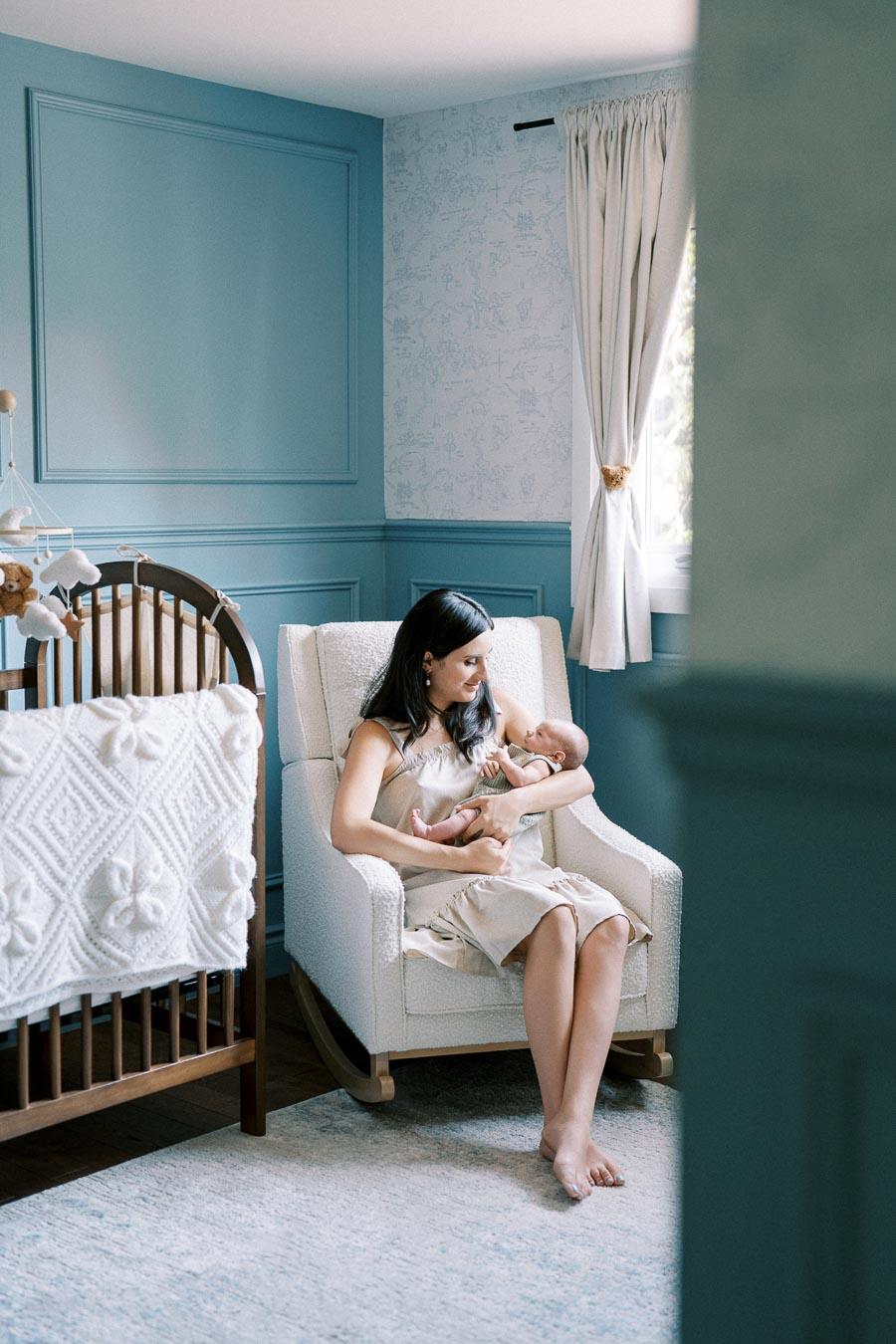 A woman gently cradling a baby in a serene, blue-themed nursery with a cozy armchair and a nearby crib adorned with a white, textured blanket.