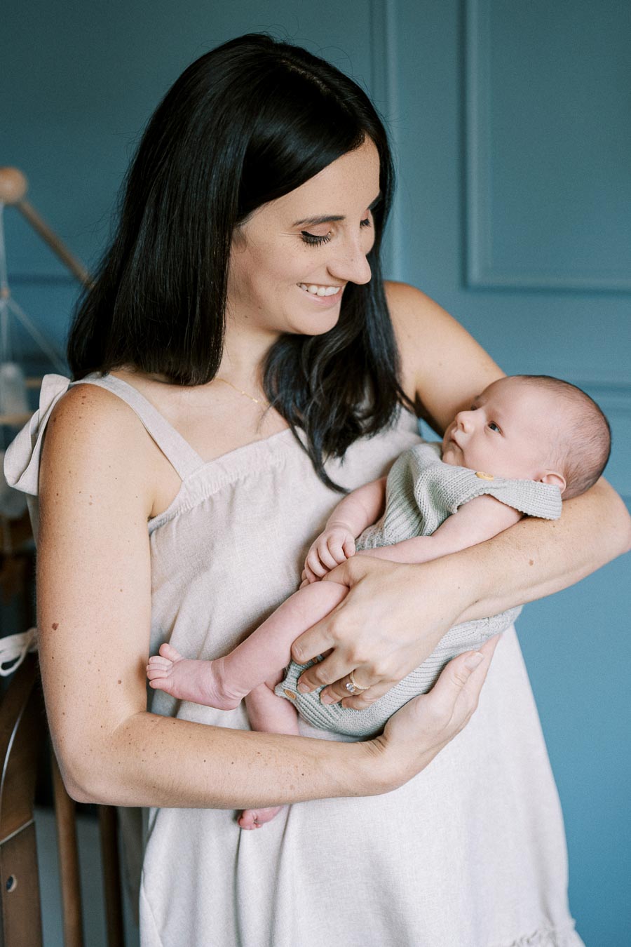 A smiling woman holding a baby in her arms, standing in a softly lit room with a blue wall.