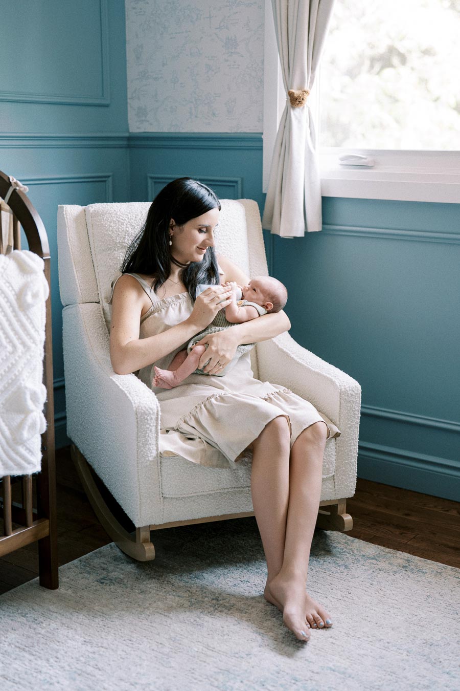 A mother sitting in a cozy nursery, gently holding and feeding her baby with a bottle while sitting on a white rocking chair.