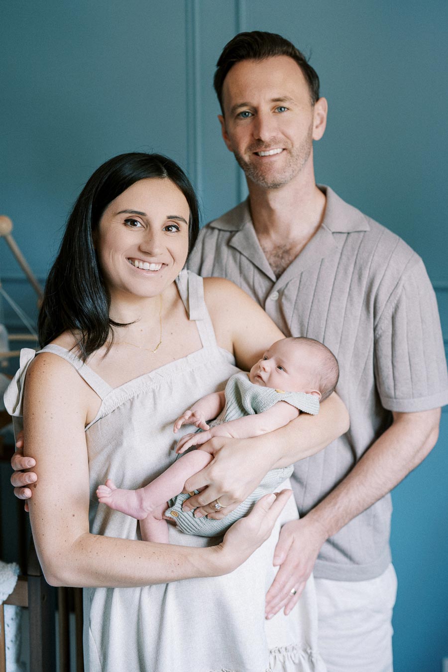 A smiling couple poses with their newborn baby, dressed in neutral tones, against a blue background in a cozy home setting.