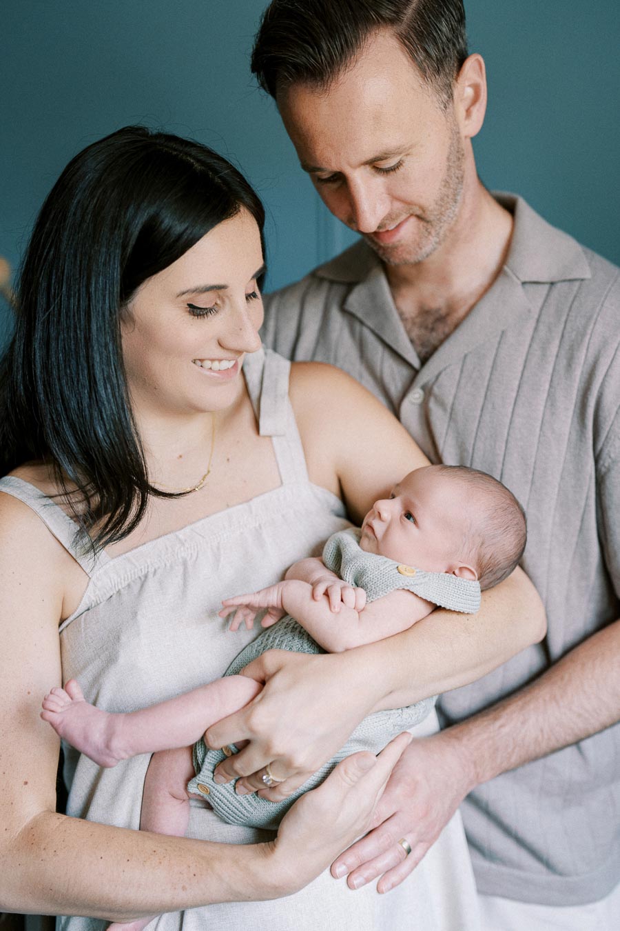 A happy couple lovingly holding their newborn baby, dressed in a knitted outfit, against a soft blue background.