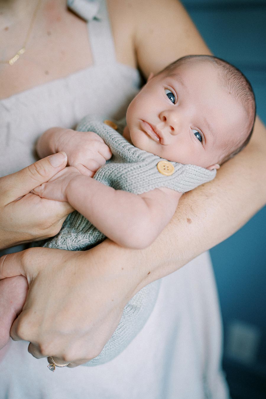 A close-up of a newborn baby with blue eyes being gently held in an adult's arms, wearing a gray knitted outfit with wooden buttons, set against a soft blue background.