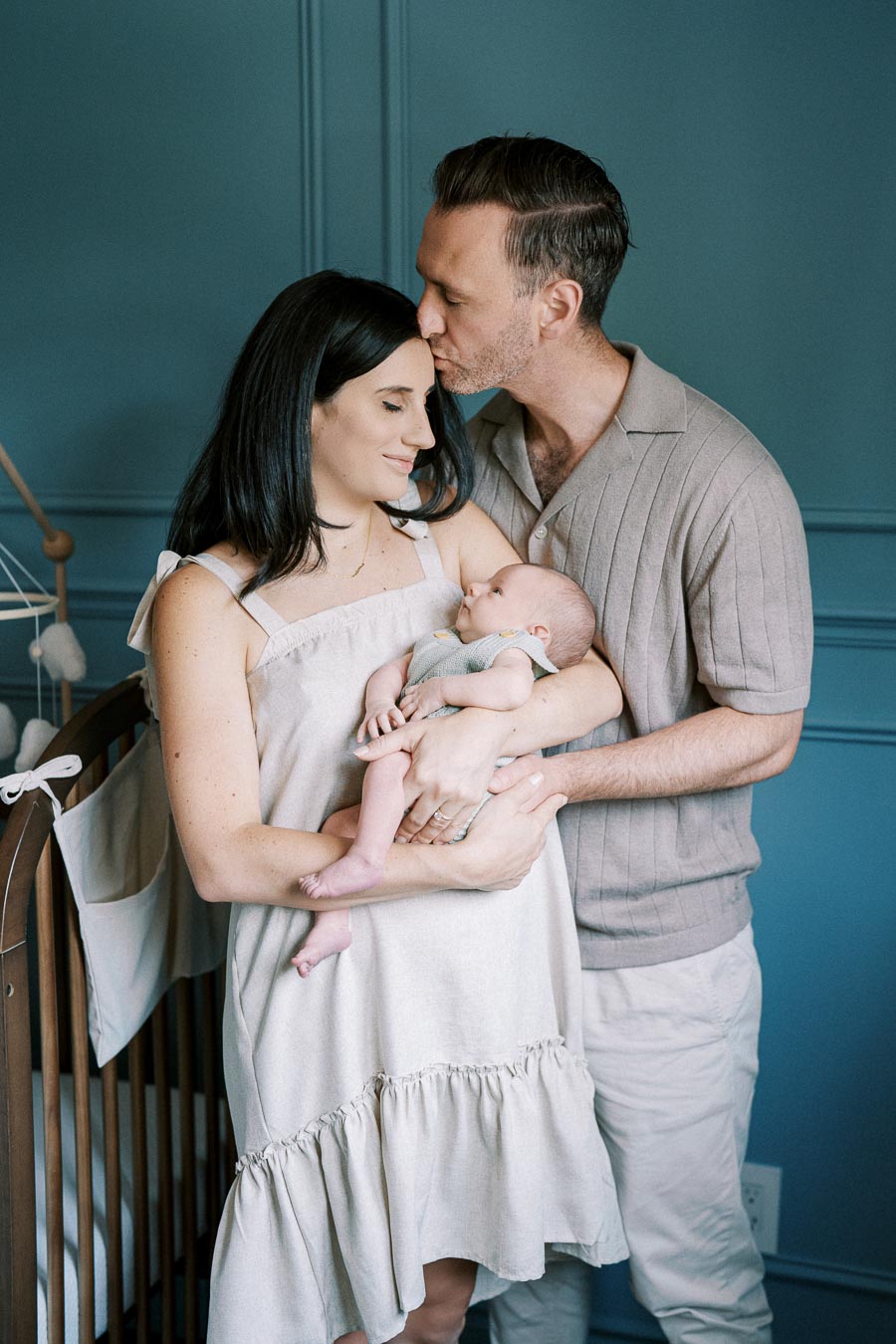 A loving couple standing in a blue nursery, holding their baby. The father gently kisses the mother's forehead as she cradles the infant in a beige dress, next to a wooden crib with a mobile.