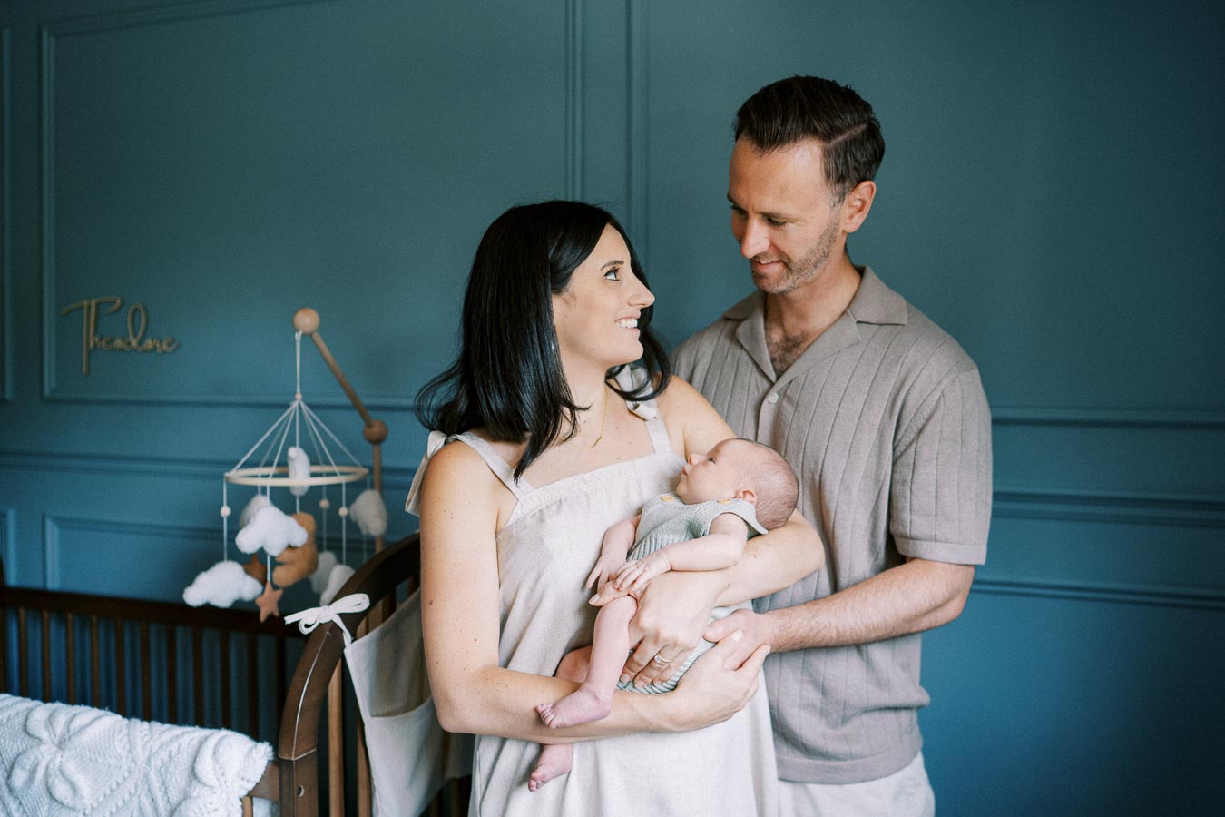 A couple lovingly gazes at their newborn baby in a serene, blue nursery with a wooden crib and cloud-themed mobile.