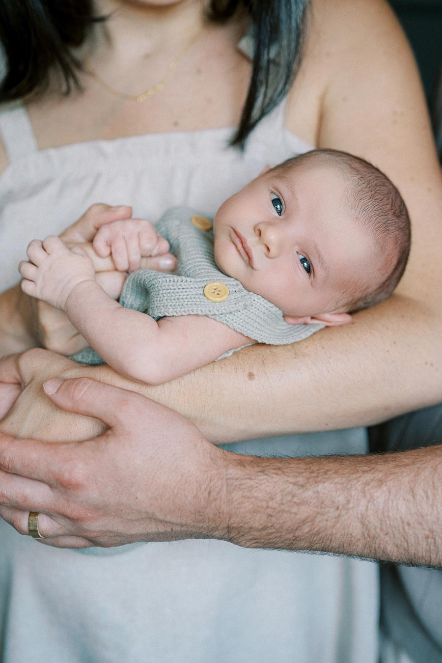 A newborn baby cradled in the arms of a parent wearing a gray sweater with wooden buttons, expressing warmth and family love.