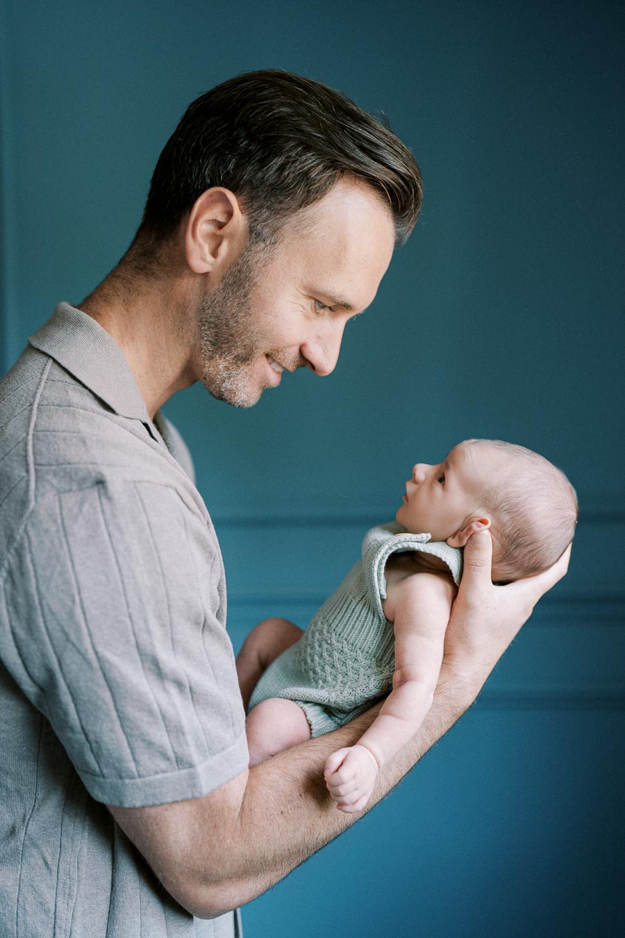 A smiling man holding a baby, gazing affectionately at each other, against a blue background.