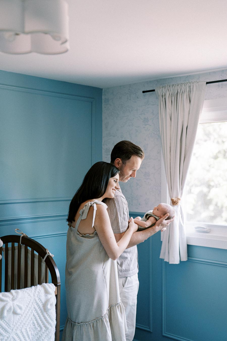 Couple lovingly holding their baby in a blue-themed nursery with natural light streaming through a window.