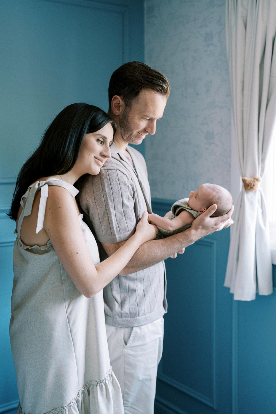 A couple lovingly gazing at their newborn baby, standing in a softly lit blue room.