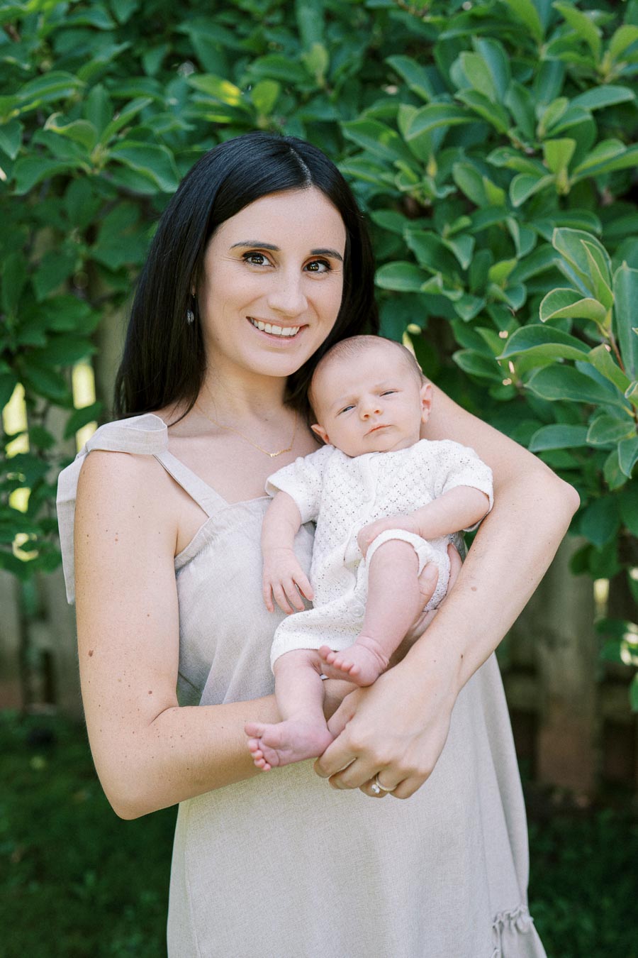 A woman holding a baby outdoors in a garden with lush green leaves in the background, both wearing light clothing, and the woman smiling at the camera.