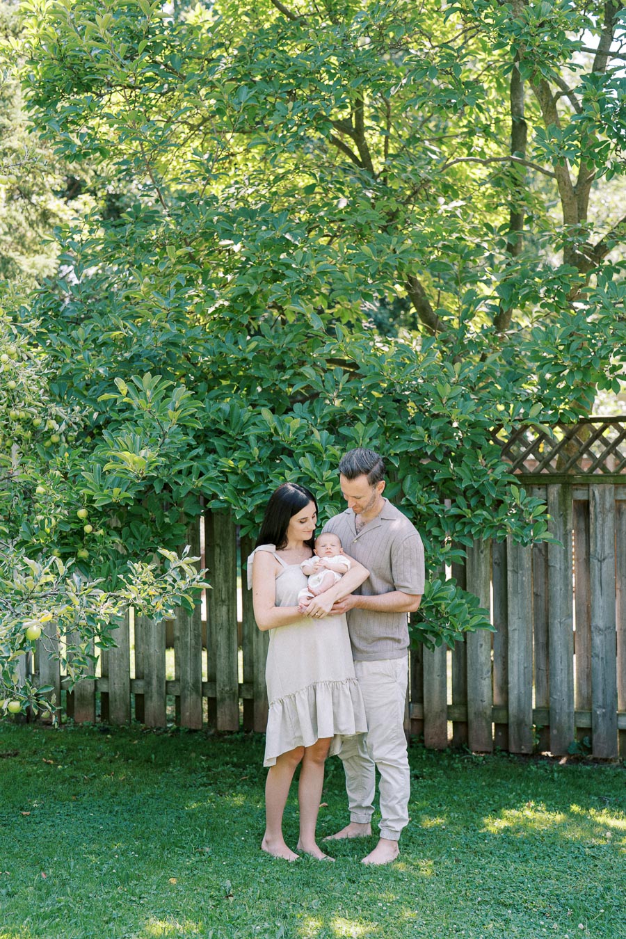 A young couple holds their baby in a lush green garden with a wooden fence and apple trees in the background.