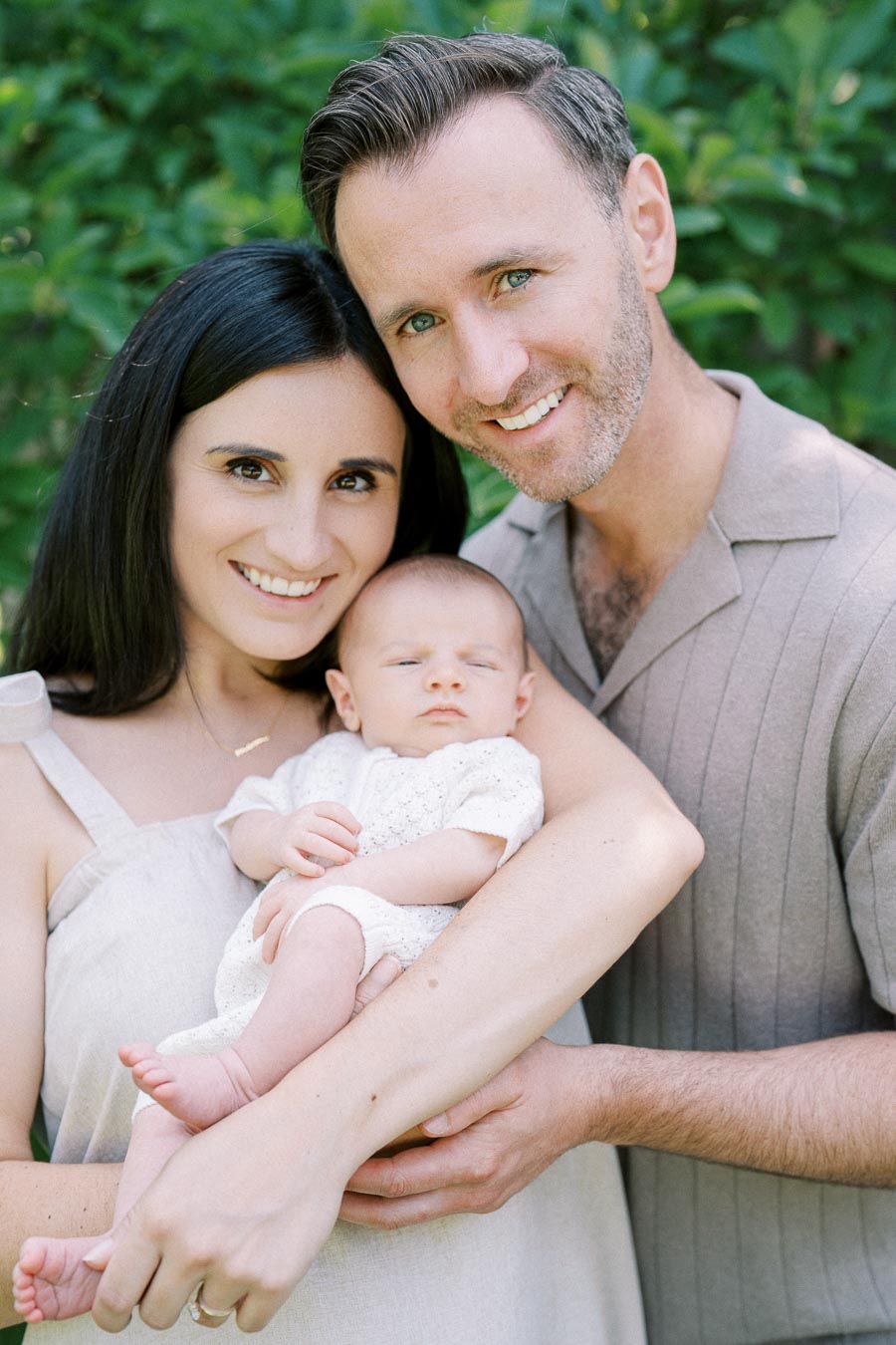 A happy couple lovingly holding their newborn baby in a lush green garden, capturing a joyful family moment.