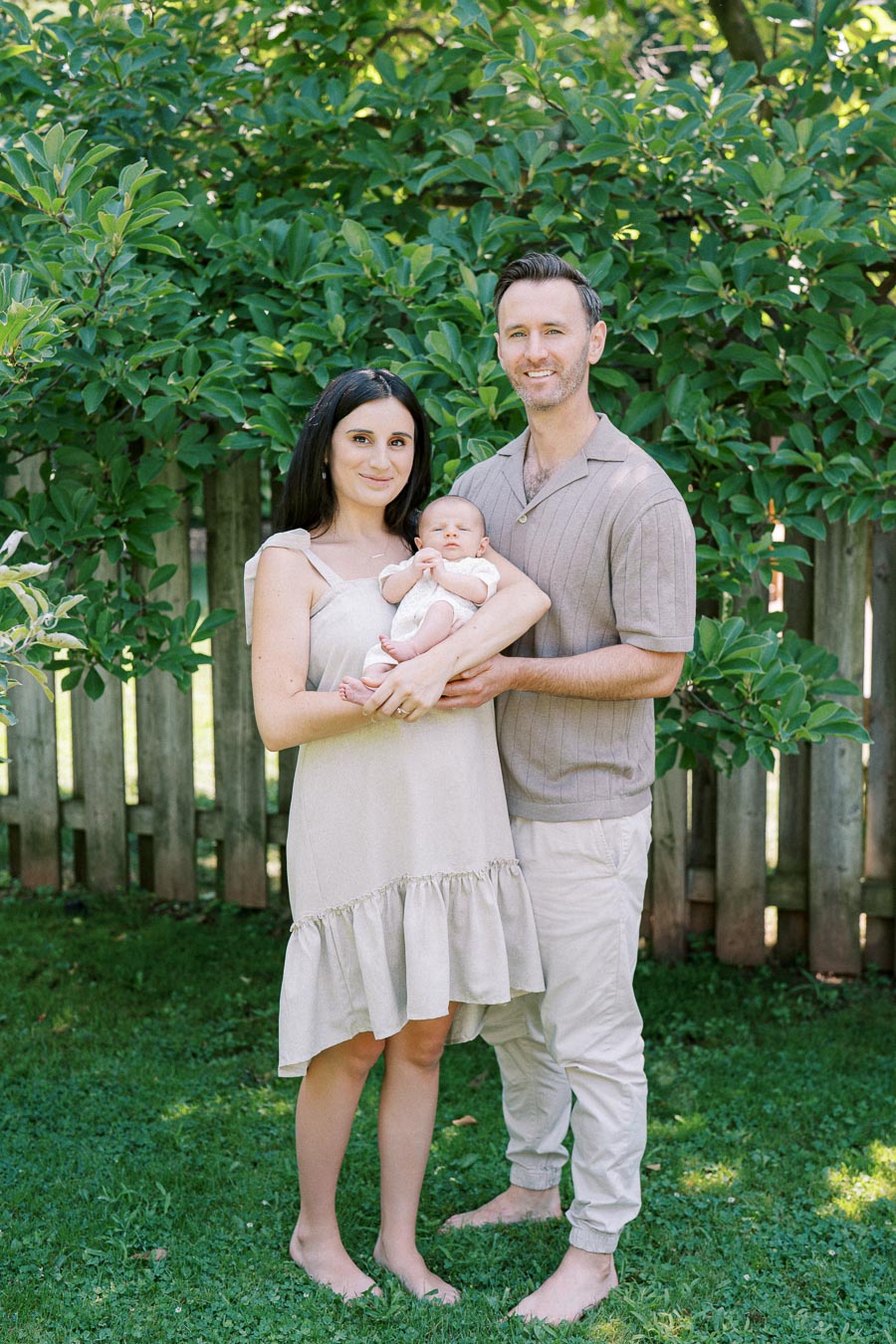 Family standing together in a garden, holding a baby, with green trees and a wooden fence in the background.