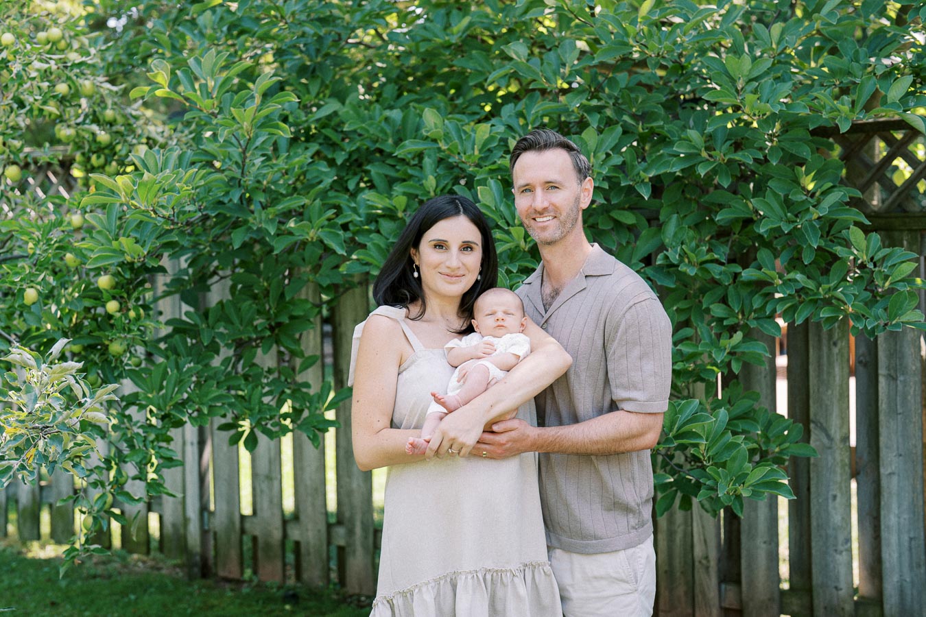 A happy family standing outdoors in a garden, with a mother and father holding their baby in front of lush green foliage and a wooden fence.