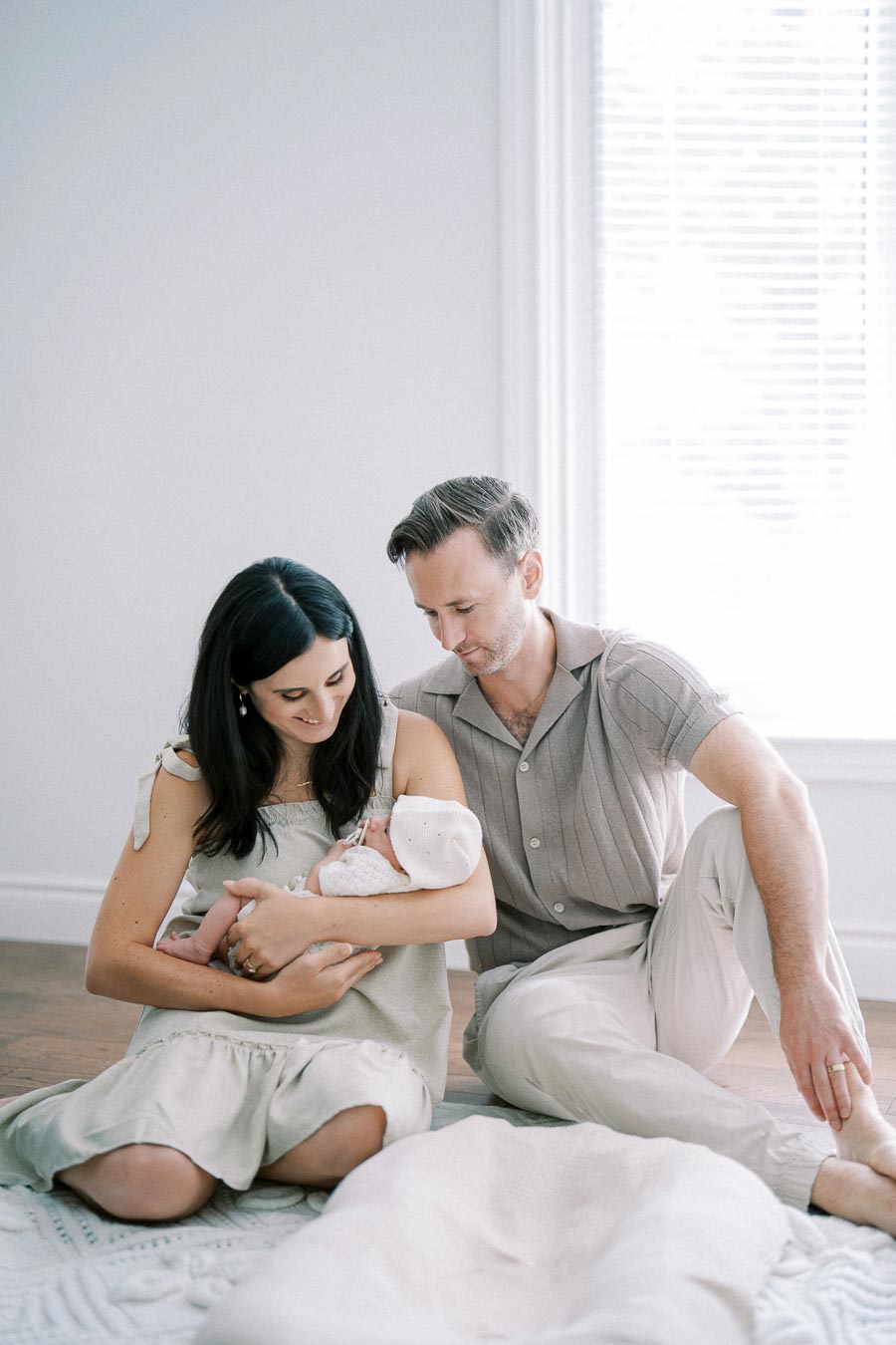 A loving couple sits on the floor in a softly lit room, gently cradling a newborn baby wrapped in a white blanket. The serene atmosphere reflects the warmth and tenderness of this family moment.