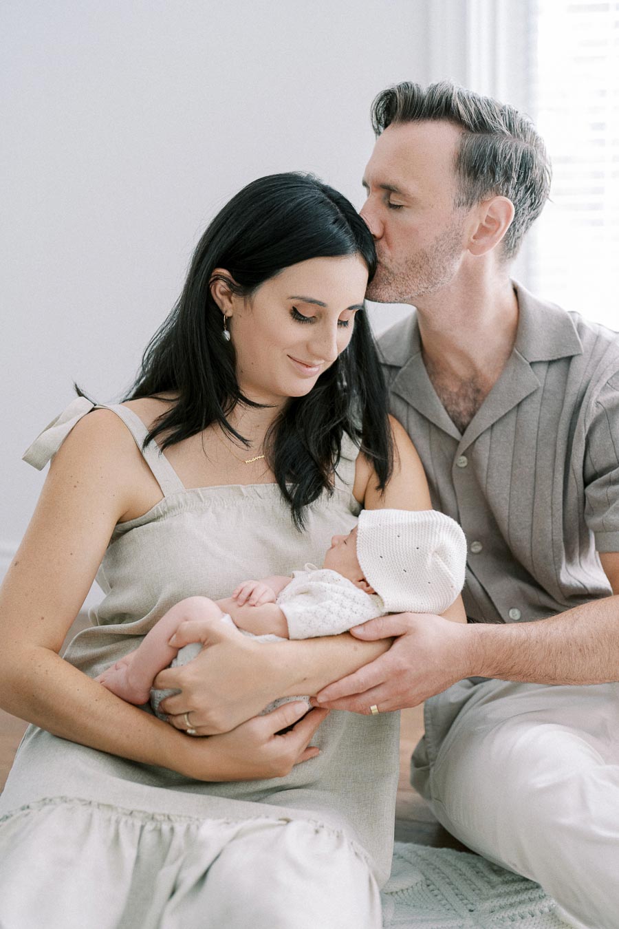 A loving couple cradles their newborn baby, the father gently kissing the mother's forehead, showcasing a tender family moment in a serene indoor setting.