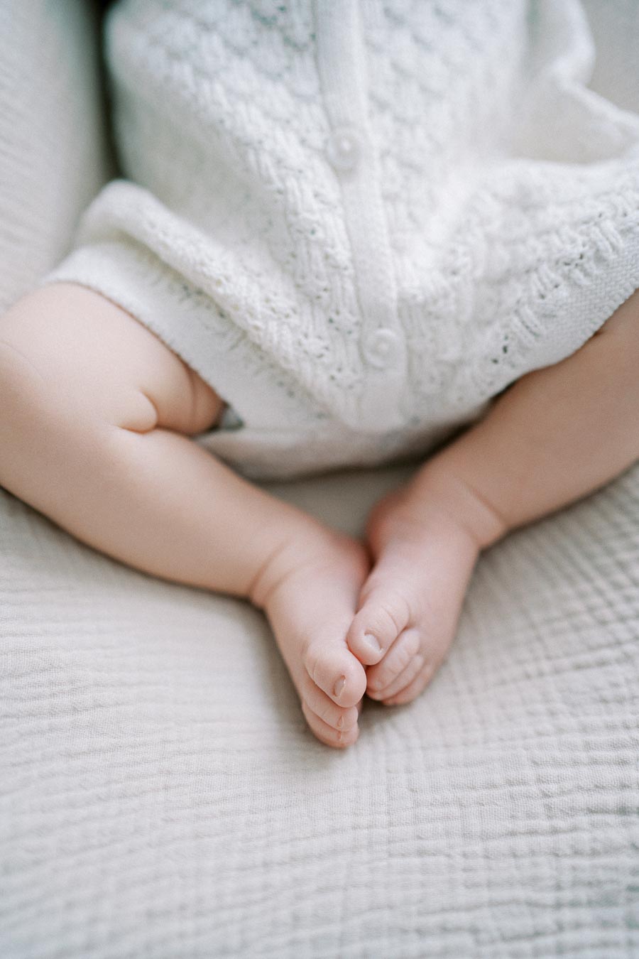Close-up of a baby's legs and feet, dressed in a soft white knitted outfit, resting on a textured neutral fabric.