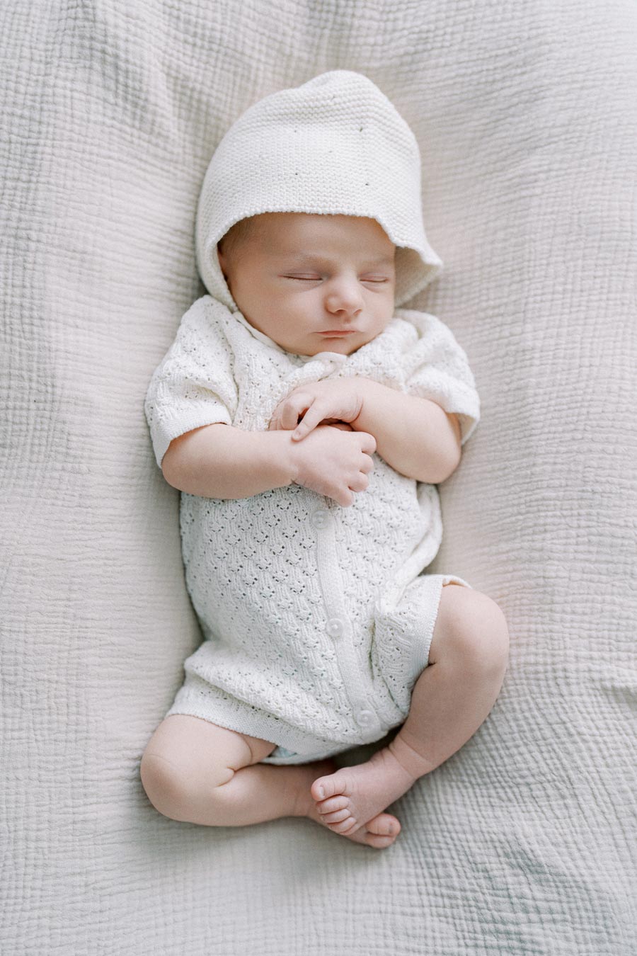Peaceful newborn baby sleeping in a white knitted outfit and hat on a soft, textured blanket.
