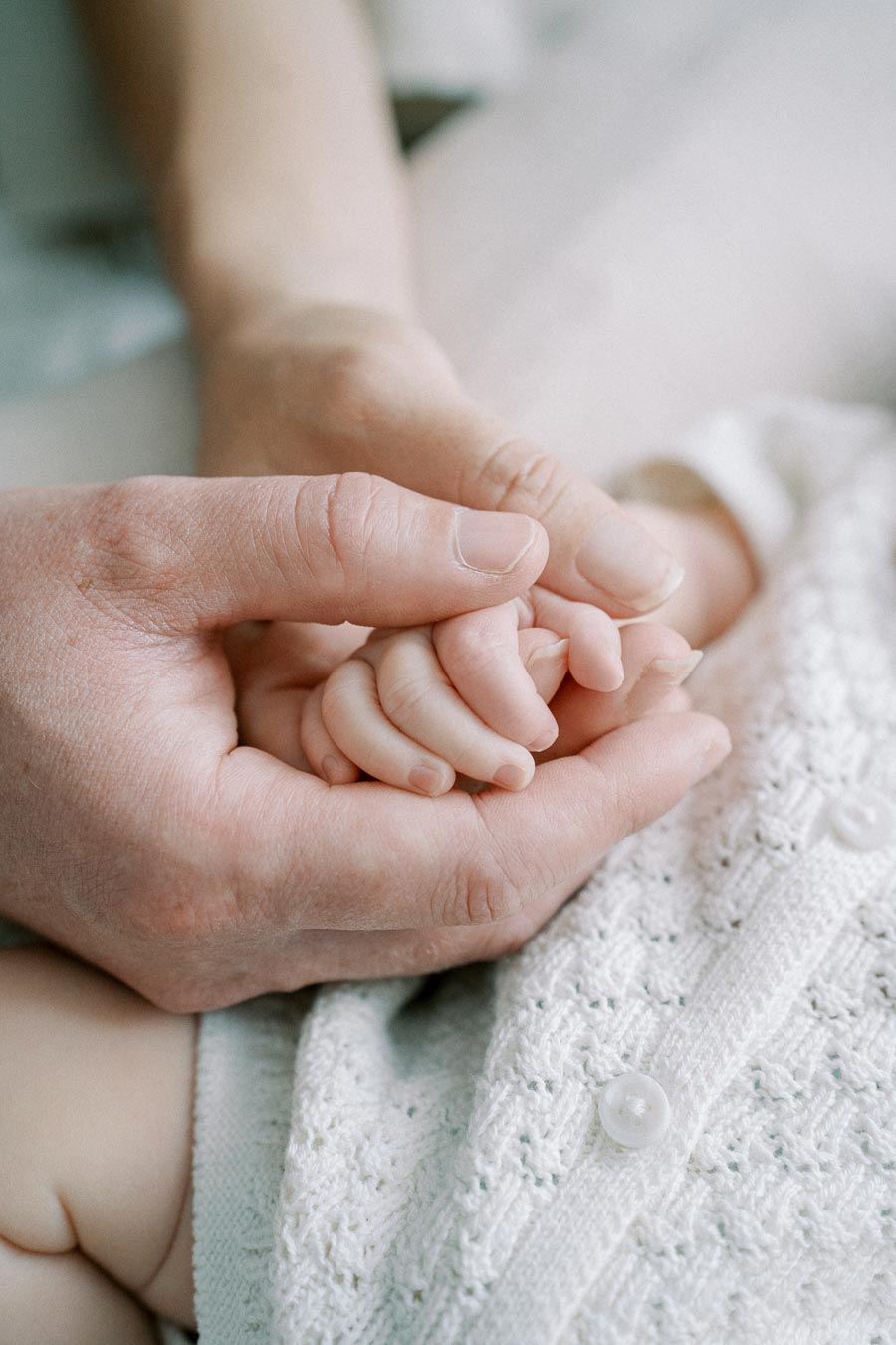 A close-up of an adult hand gently holding a baby's hand, symbolizing care, love, and family bonds, with a soft focus on the knitted fabric in the background.