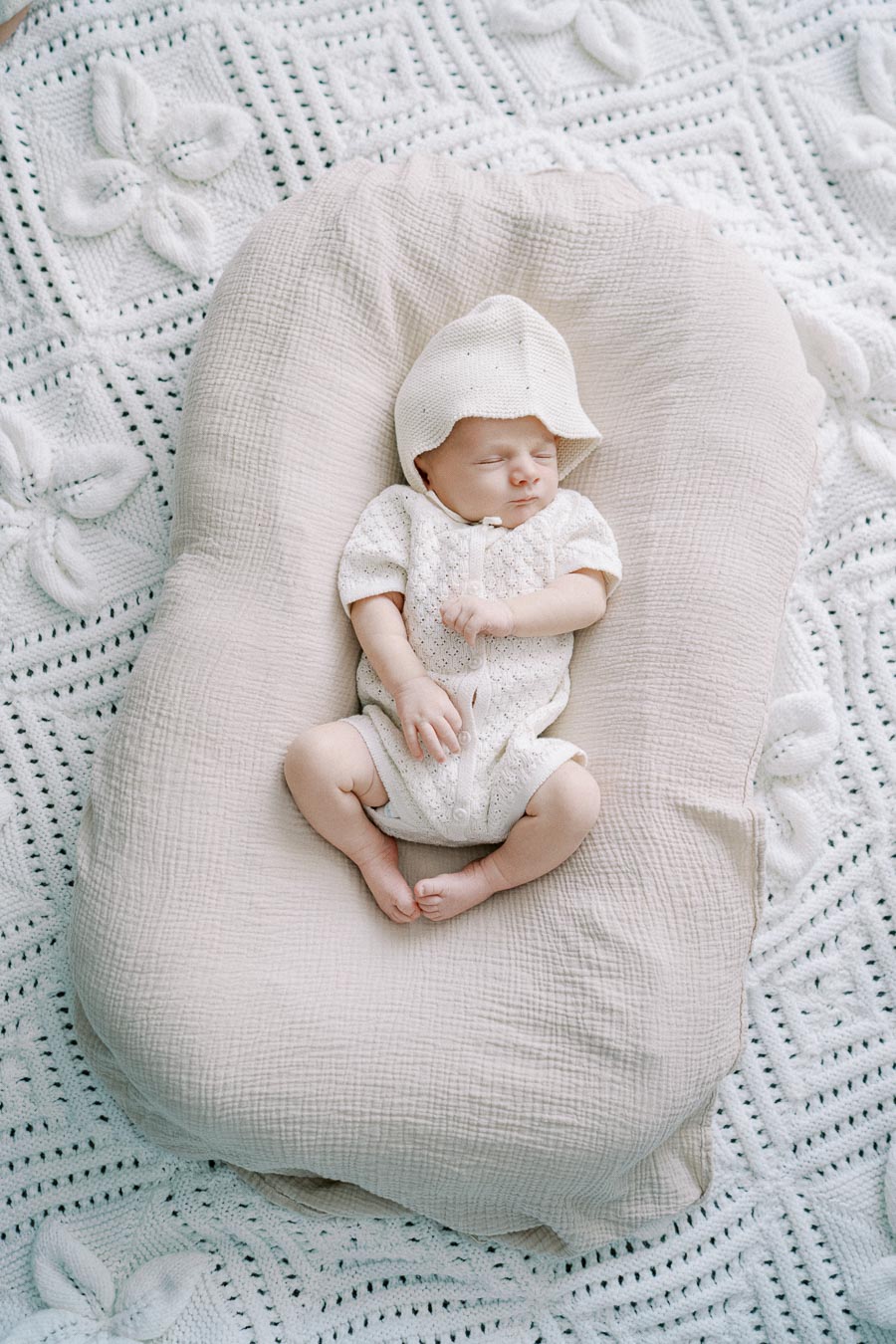Sleeping newborn baby in a white knitted outfit and hat, resting on a soft beige cushion with a crocheted blanket background.