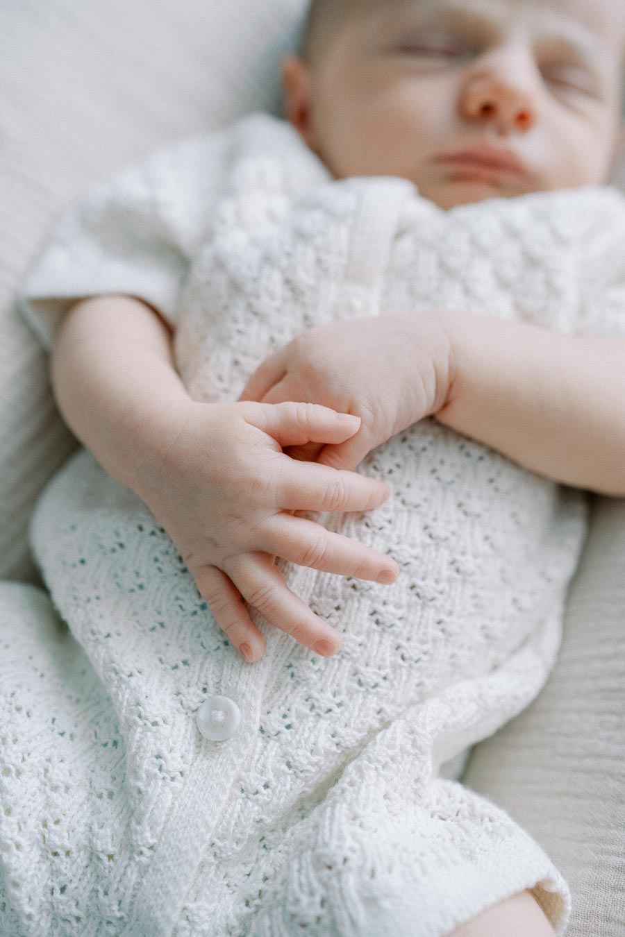 Sleeping newborn baby wearing a white knitted outfit, with hands gently clasped on a soft blanket.