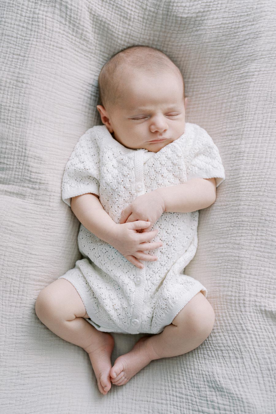 Peaceful sleeping baby wearing a white knitted onesie, lying on a soft, textured blanket.