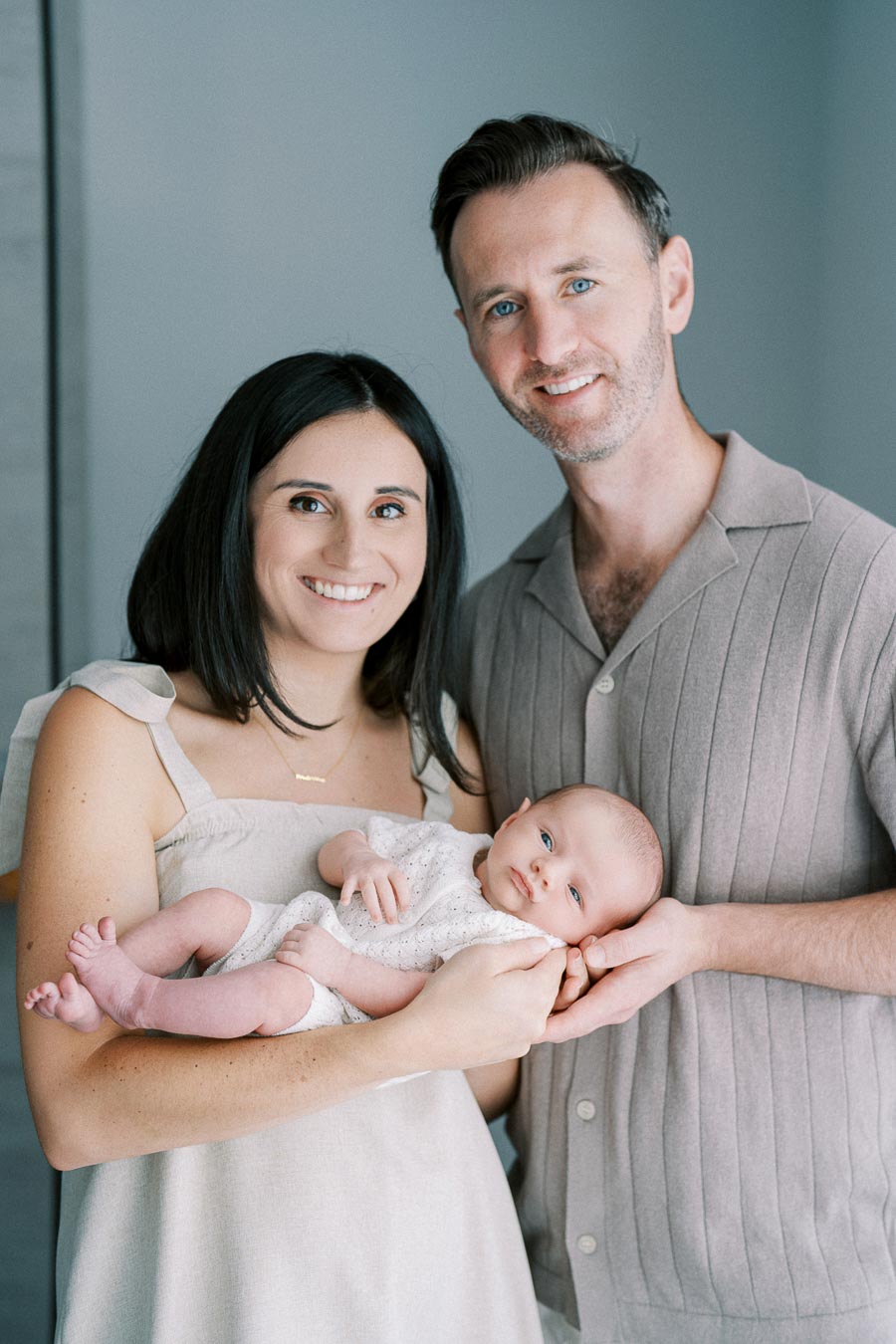 Friendly family of three posing together, with the mother holding a baby and the father standing close, all smiling warmly.