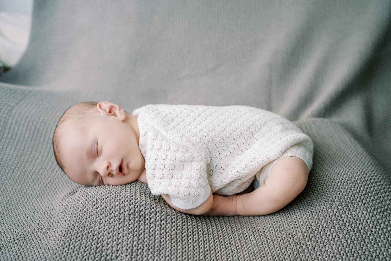 Peaceful newborn baby sleeping on a soft, gray knitted blanket, wearing a white, textured onesie.
