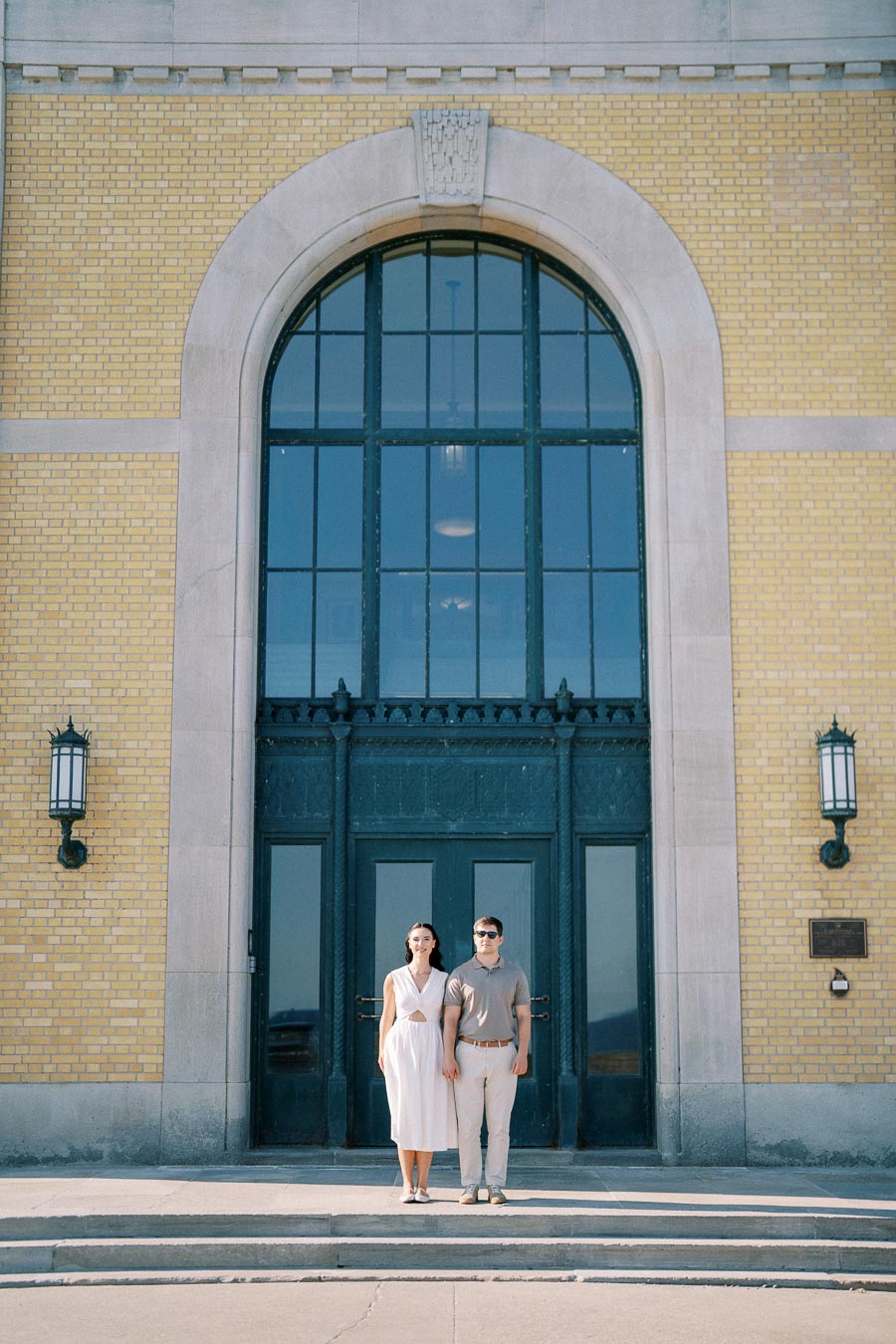 A couple standing in front of a grand, arched doorway with yellow brick walls, holding hands on a sunny day.
