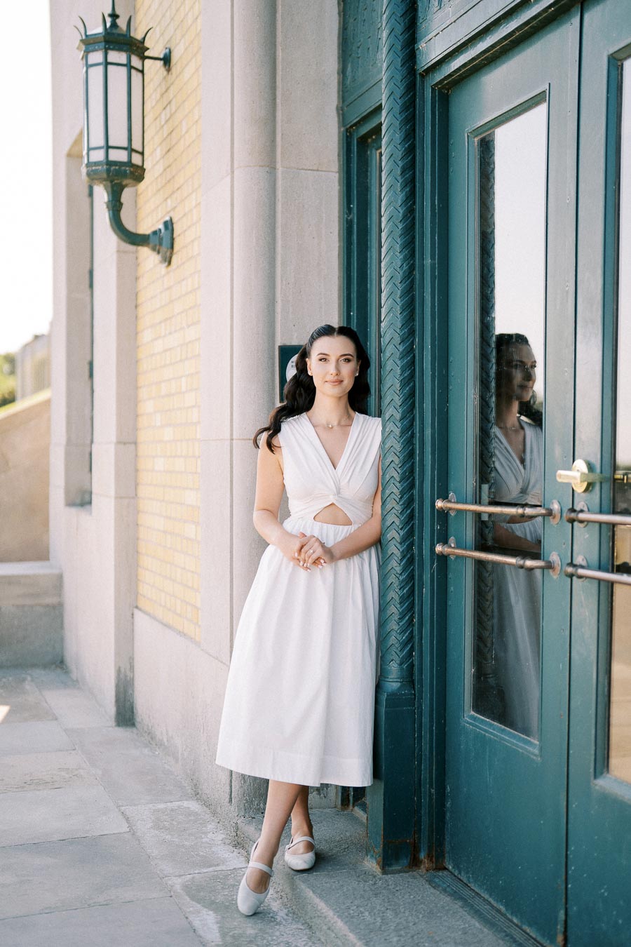 A woman in a stylish white dress stands leaning against a green door, with vintage outdoor lighting and yellow brick wall in the background.