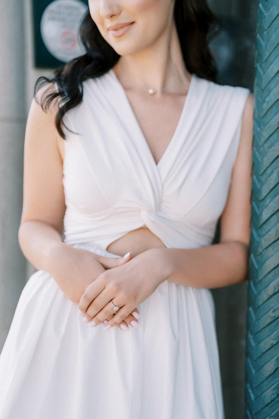 Elegant woman in a stylish white dress showcasing a diamond engagement ring, standing beside a textured wall.