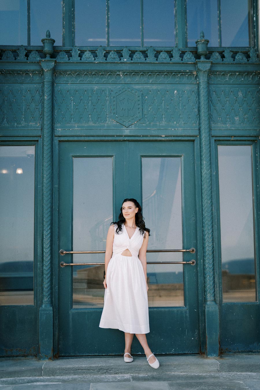 A woman in a white dress standing in front of a large, ornate teal door with intricate designs.