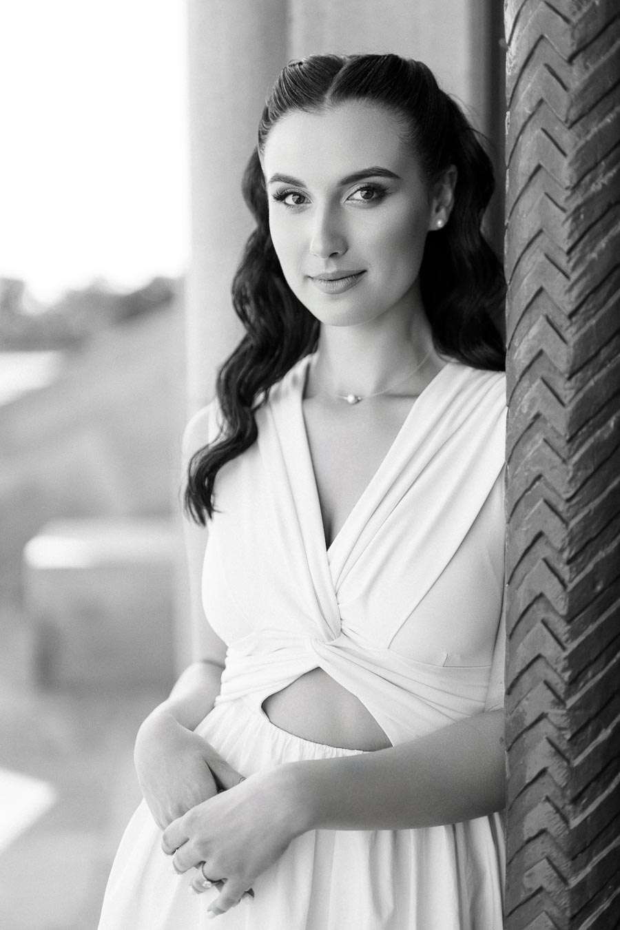 Black and white portrait of a woman in an elegant white dress, standing against a patterned wall, looking confidently into the camera.