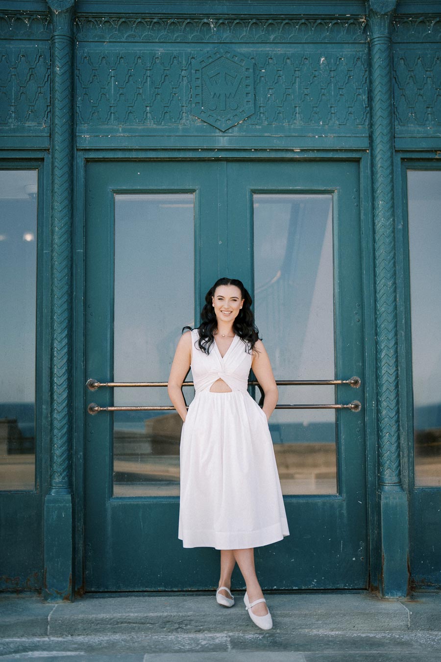 A woman in a white dress stands in front of ornate double doors, smiling confidently, with a detailed, green geometric design above her.